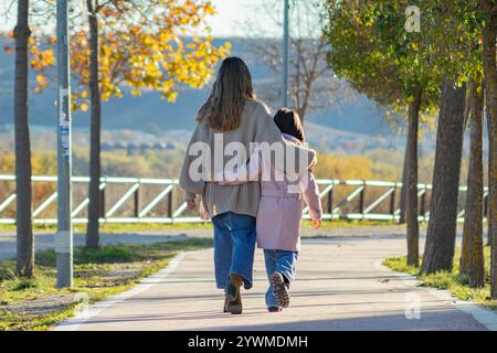 Eine herzerwärmende Szene, in der eine Mutter und ihre Tochter zusammen, umarmt, einen friedlichen Pfad entlang gehen, umgeben von Herbstbäumen. Beide sind in Coz gekleidet Stockfoto