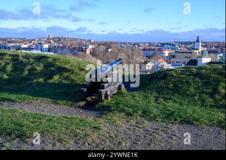 Eine historische Kanone befindet sich auf grasbewachsenem Gelände und bietet einen atemberaubenden Blick auf die Skyline der Stadt unter einem klaren blauen Himmel. Die Szene erfasst eine Mischung aus n Stockfoto