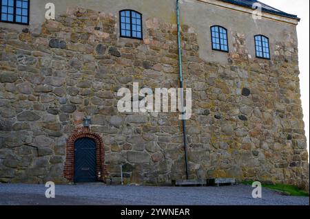 Eine stabile Steinmauer verfügt über große, unregelmäßige Steine und mehrere Fenster. Eine bogenförmige Holztür verleiht Charme, in einer reichen historischen Umgebung gelegen Stockfoto