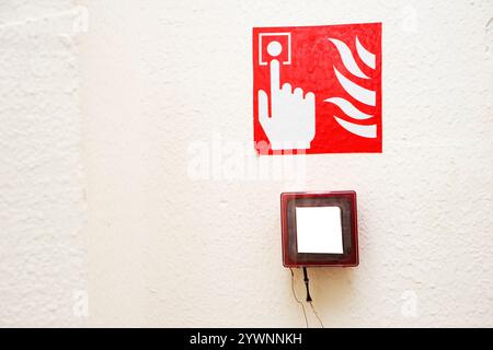 Feueralarm-Taste, die in Glas über einem Schild mit einem Finger in der Nähe von Flammen an einer weißen Wand eingehängt ist Stockfoto