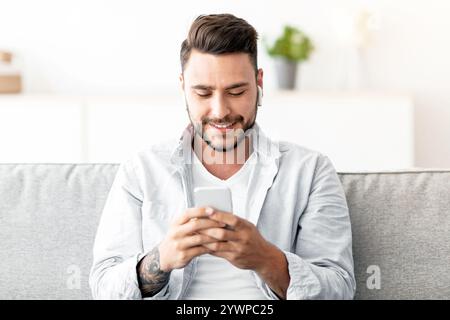 Junger Mann in Ohrhörern, der auf dem Smartphone chattet und zu Hause auf dem Sofa sitzt, freier Platz. Technologie und Gadgets Konzept Stockfoto