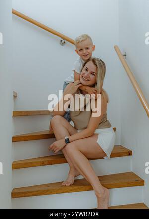 Glückliche Mutter und dreijähriger Sohn sitzen auf der Treppe in ihrem Haus mit weißen Wänden an den Seiten des Hauses. Hochwertige Fotos Stockfoto