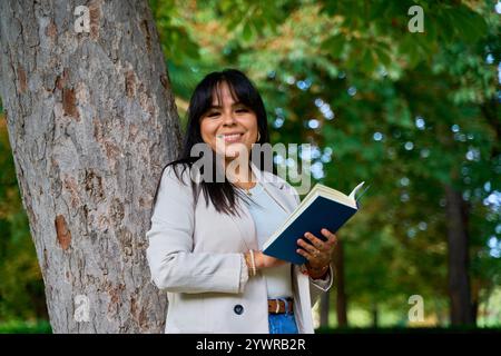 Latina liest ein Buch, während sie sich an einen Baum im Park lehnt Stockfoto