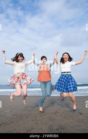 Drei junge Frauen springen auf den Sandstrand und halten die Hände Stockfoto