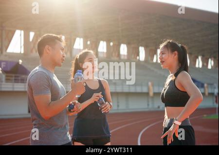 Eine Gruppe asiatischer Läufer genießt nach dem Training in einem Stadion an einem hellen, sonnigen Tag eine Unterhaltung, während sie sich auf einer Laufstrecke ausruhen. Personen und Stockfoto