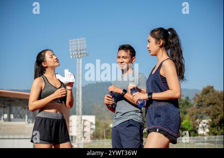 Eine Gruppe asiatischer Läufer genießt nach dem Training in einem Stadion an einem hellen, sonnigen Tag eine Unterhaltung, während sie sich auf einer Laufstrecke ausruhen. Personen und Stockfoto