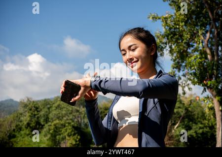 Eine schöne, sportliche asiatische Frau in Sportbekleidung überprüft ihre Kalorienanzahl auf ihrer Smartwatch, während sie an einem Sommertag in einem Park läuft. Sport-Gadget, Lifest Stockfoto