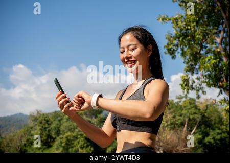 Eine schöne, sportliche asiatische Frau in Sportbekleidung überprüft ihre Kalorienanzahl auf ihrer Smartwatch, während sie an einem Sommertag in einem Park läuft. Sport-Gadget, Lifest Stockfoto