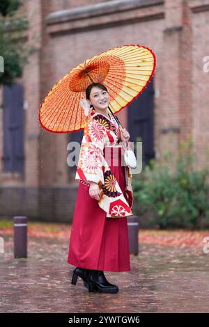 Eine Japanerin im Alter von 20 Jahren, die einen roten Kimono (Hakama) trägt, ein Grundnahrungsmittel japanischer Universitätsabsolventen, lächelt und posiert im Regen mit einem japanischen um Stockfoto