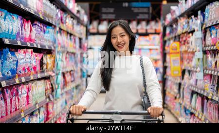 Chiang Mai, Thailand - 09. Dezember 2024: Eine attraktive, lächelnde asiatische Frau, die in einem Supermarkt einkauft, einen Einkaufswagen schiebt, durch einen Gang läuft, A Stockfoto