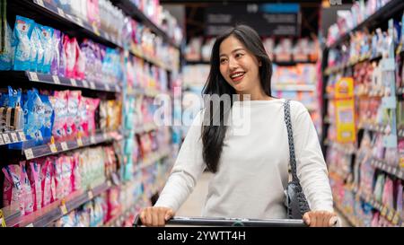Chiang Mai, Thailand - Dez 09 2024: Eine attraktive, glückliche asiatische Frau, die in einem Supermarkt einkaufen, einen Einkaufswagen schieben, durch einen Gang gehen und Stockfoto
