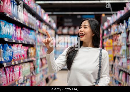 Chiang Mai, Thailand - 09. Dezember 2024: Eine attraktive, glückliche asiatische Frau, die in einem Supermarkt einkaufen und mit dem Finger nach Artikeln auf einer sie sucht Stockfoto