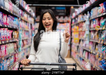 Chiang Mai, Thailand - Dez 09 2024: Eine schöne asiatische Frau, die in einem Supermarkt einkauft, mit einem Einkaufswagen in einem Gang steht und den Camer anlächelt Stockfoto