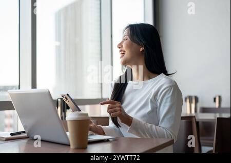 Eine fröhliche, positive asiatische Frau, die lacht, während sie Nachrichten auf ihrem Smartphone liest, aus der Ferne in einem Café in einem modernen Gebäude arbeitet, sitzt Stockfoto