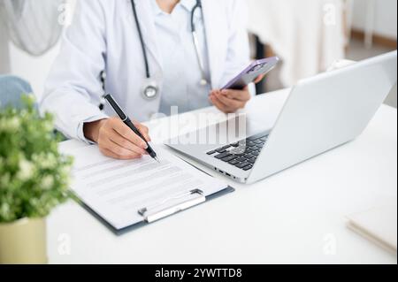 Ein zugeschnittenes Bild einer Ärztin, die an ihrem Schreibtisch im Büro arbeitet, ihr Smartphone hält, während sie medizinische Papiere liest, vor einem Computer sitzt. Stockfoto