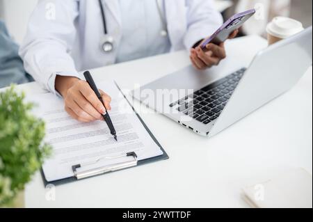 Ein zugeschnittenes Bild einer Ärztin, die an ihrem Schreibtisch im Büro arbeitet, ihr Smartphone hält, während sie medizinische Papiere liest, vor einem Computer sitzt. Stockfoto