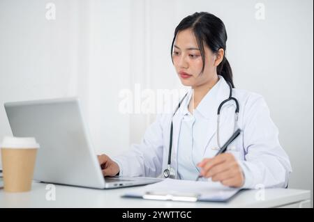 Eine attraktive asiatische Ärztin in Uniform, die sich auf das Lesen von medizinischen Fällen in Papier und auf ihrem Laptop konzentriert und an ihrem Schreibtisch im Büro des arbeitet Stockfoto