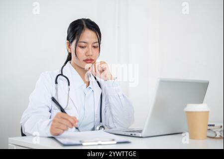 Eine attraktive asiatische Ärztin in Uniform, die sich auf das Lesen von medizinischen Fällen in Papier und auf ihrem Laptop konzentriert und an ihrem Schreibtisch im Büro des arbeitet Stockfoto