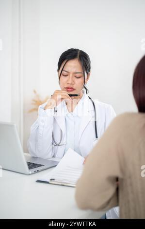 Eine ernsthafte, gestresste asiatische Ärztin, die während einer ärztlichen Konsultation im Untersuchungsraum des hospits aufmerksam auf die Symptome eines Patienten hört Stockfoto