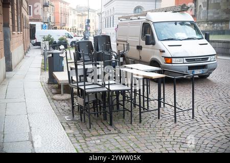 Cremona, Italien - 27. November 2024 gestapelte Stühle und Tische überladen eine kopfsteingepflasterte Straße, in der Nähe parkt ein weißer iveco Daily Van, was auf ong hindeutet Stockfoto