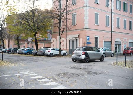 Cremona, Italien - 27. November 2024 Silver Smart für zwei elektrische Antriebszweige, die an einer Kreuzung in einer städtischen Straße mit geparkten Autos und einem rosafarbenen Gebäude abbiegen Stockfoto