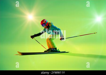 Leuchtende Grüntöne versetzen den Skifahrer in Bewegung, ihr farbenfrohes Outfit leuchtet vor dem beleuchteten Gradient-Studio-Hintergrund. Stockfoto
