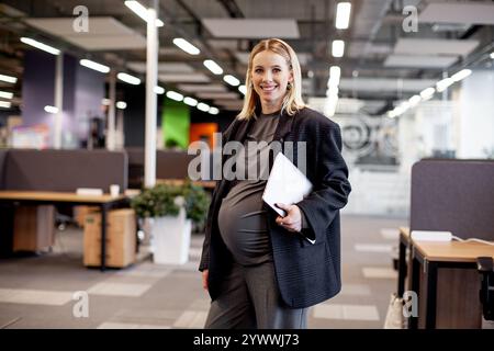Selbstbewusste schwangere Geschäftsfrau, die in einem modernen Büro steht, ein Tablet hält und lächelt und das Gleichgewicht zwischen Karriere und Mutterschaft in einem profi zeigt Stockfoto