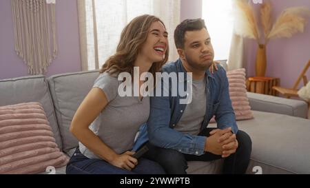 Frau lacht, während der Mann auf einer Couch in einem gemütlichen Wohnzimmer langweilig aussieht, was verschiedene Emotionen in der intimen Umgebung eines Paares suggeriert. Stockfoto