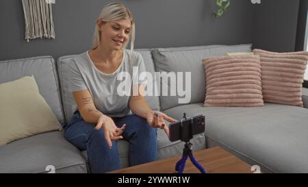 Junge Frau mit blonden Haaren, die drinnen auf einer Couch sitzt, sich zu Hause im Wohnzimmer mit einem Smartphone auf einem Stativ aufnimmt, lächelnd und gestu Stockfoto