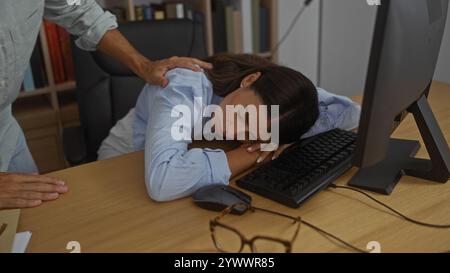 Frau schläft an ihrem Schreibtisch in einem Büro, während ein Kollege sie sanft weckt, umgeben von einem Computer, einer Tastatur und einer Brille Stockfoto