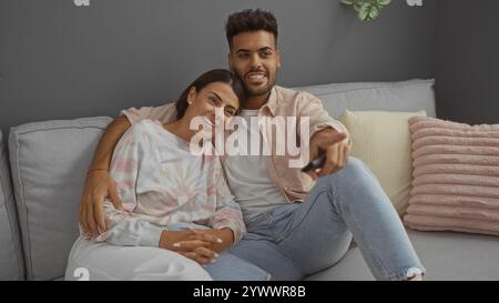 Paare entspannen auf einer gemütlichen Couch in einem Wohnzimmer, während der Mann auf den Fernseher zeigt, während er eine Fernbedienung hält, um einen Moment der Freizeit zu teilen Stockfoto