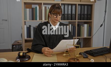 Frau Richterin, die in einem Büro arbeitet und Dokumente an ihrem Schreibtisch liest, umgeben von Gesetzbüchern und juristischen Symbolen, wie einem Hammer und einer Waage der Gerechtigkeit. Stockfoto
