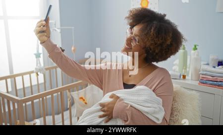Frau mit lockigen Haaren hält Ihr Baby im Schlafzimmer lächelnd, während Sie Selfie in gemütlichem Wohnraum machen, was die fröhliche Mutterschaft widerspiegelt. Stockfoto