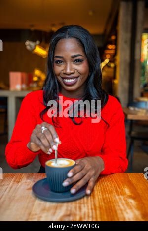 Lächelnde schwarze Frau rührt Cappuccino im Café mit rotem Pullover Stockfoto