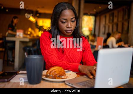Junge Frau, die im Café an einem Laptop arbeitet und Kaffee und Gebäck genießt Stockfoto