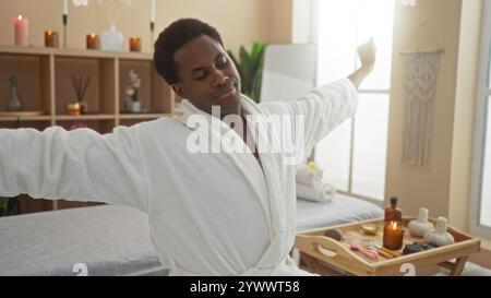 Junger Mann, der sich in einem Spa in einem weißen Bademantel ausdehnt, umgeben von Kerzen und Wellness-Produkten in einem ruhigen Schönheitsraum Stockfoto