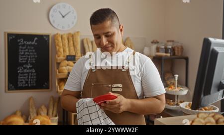 Junger Mann, der eine Schürze trägt, lächelt beim Putzen in der Bäckerei mit Brotregalen im Hintergrund Stockfoto