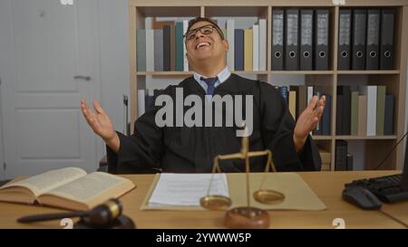 Junger hispanischer Mann, als Richter verkleidet, sitzt in einem Büro mit Gesetzbüchern und Waagen auf dem Tisch und drückt Freude aus Stockfoto