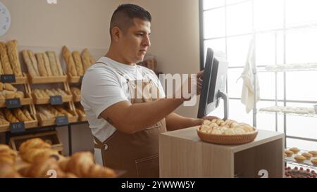 Lateinischer Mann, der in einer Bäckerei arbeitet, mit einem digitalen Bildschirm, um Bestellungen in einem Innenbereich zu verwalten, mit Regalen voller frischem Brot und Gebäck. Stockfoto