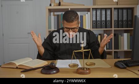 Junger hispanischer Mann in einem Richtermantel in einem Büro, umgeben von Rechtsbüchern, Waagen der Gerechtigkeit und einem Hammer auf dem Schreibtisch, in einem gerichtlichen indo Stockfoto