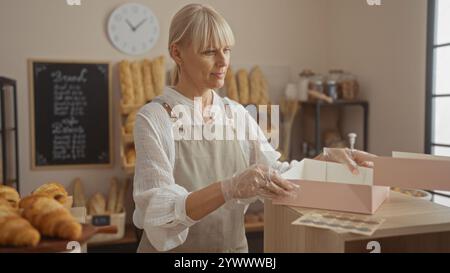 Frau, die Bäckerei zubereitete, blonder reifer Erwachsener mit Handschuhen im Ladeninneren Stockfoto