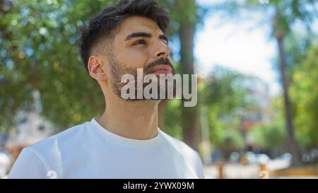 Junger hispanischer Mann mit Bart, der an einem sonnigen Tag draußen in einem urbanen Park steht Stockfoto