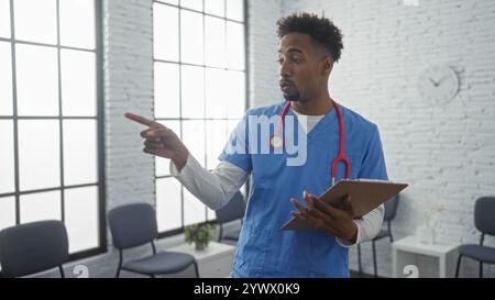 Eine junge, gutaussehende afroamerikanische Krankenschwester mit Bart hält ein Klemmbrett und zeigt in einem hellen Wartezimmer des Krankenhauses. Stockfoto