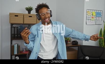 Ein junger afroamerikanischer Mann, der Musik mit Kopfhörern genießt, ein Smartphone hält und begeistert in einem modernen Büro tanzt. Stockfoto