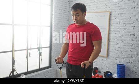 Junger chinesischer Mann, der in einem Fitnessraum in einem gut beleuchteten Sportzentrum mit Geräten wie Widerstandsbändern und einem Whiteboard trainiert. Stockfoto