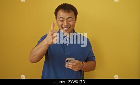Ein hübscher junger chinese in einem blauen Hemd lächelt und zeigt einen Daumen hoch, während er ein Smartphone hält, vor einem isolierten gelben Hintergrund W Stockfoto