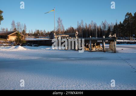 Ein ruhiger Wintermorgen in Leksand zeigt ein hölzernes Dock, das sich über einen gefrorenen See erstreckt, umgeben von Schnee und Bäumen. Ein klarer blauer Himmel trägt zur Ruhe bei Stockfoto