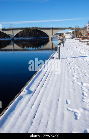 Ein ruhiger Wintertag in Leksand, Dalarna, bietet einen schneebedeckten Fußweg neben einem ruhigen Fluss. Fußspuren führen entlang des Weges, während eine Brücke Th überspannt Stockfoto