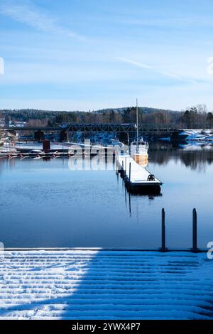 Schnee bedeckt die Ufer einer ruhigen Uferpromenade in Leksand, Dalarna. Boote ruhen friedlich am Dock unter einem klaren blauen Himmel. Eine ruhige Winterszene Stockfoto