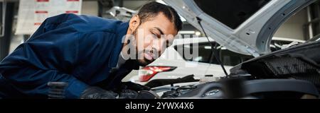 Ein junger Mechaniker inspiziert den Motor eines Autos in einer lebhaften Autowerkstatt. Stockfoto